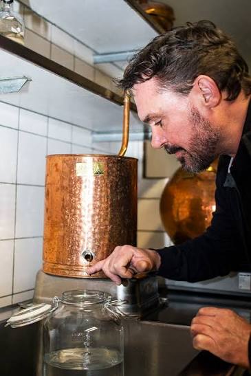 Henrik Hammer beside a copper still, drawing a sample into a glass jar.